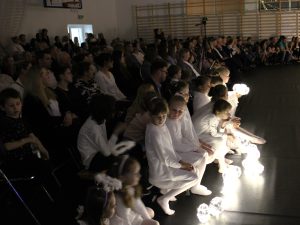 Widownia wypełniona dorosłymi i dziećmi obserwuje występ. Na pierwszym planie dzieci w białych strojach siedzące na brzegu sceny trzymają świecące kuliste rekwizyty.