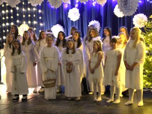 Dzieci w białych strojach aniołów stoją na scenie i śpiewają. W tle znajdują się świąteczne dekoracje z gwiezdnymi lampkami i papierowymi ozdobami.