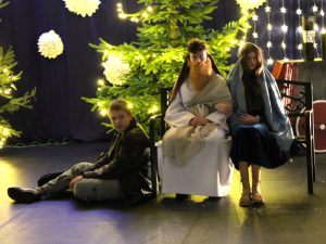 Dzieci odgrywające postaci Józefa i Maryi siedzą na ławce, a obok nich na podłodze siedzi chłopiec w ciemnym stroju. Za nimi znajdują się ozdobione choinki i światełka