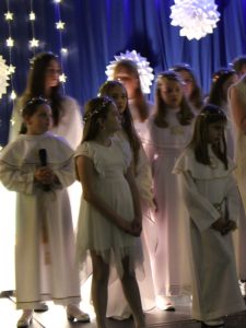 Zbliżenie na grupę dzieci w białych strojach aniołów stojących na scenie. W tle widoczne gwiezdne światełka i papierowe ozdoby.