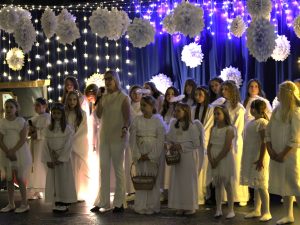 Grupa dzieci i młodzieży w białych strojach aniołów stoi na scenie. Wśród nich stoi nauczycielka, która śpiewa do mikrofonu. W tle wiszące papierowe ozdoby i światełka tworzące świąteczną scenerię.