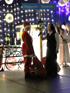 Uczestnik przedstawienia w stroju Mikołaja stoi na scenie z dużym czerwonym workiem. Obok stoi dziecko w ciemnej kamizelce, trzymające mikrofon. Za nimi dekoracja z tabliczką 'Ostaszewo Toruńskie’