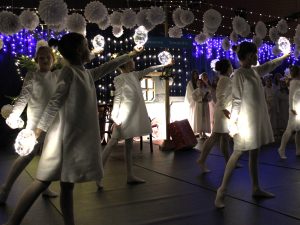 Grupa dzieci w białych strojach wykonuje taniec, trzymając w dłoniach świecące kuliste rekwizyty. W tle dekoracje świąteczne oraz grupa uczniów w strojach aniołów.