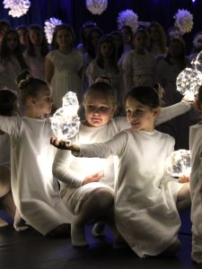 Dzieci w białych kostiumach klęczą i trzymają świecące kule. Za nimi w półcieniu stoi grupa uczniów w białych strojach aniołów.