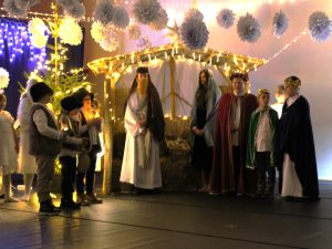 Dzieci odgrywają scenę Bożego Narodzenia. Uczestnicy w strojach Maryi, Józefa, Trzech Króli i pasterzy stoją przed szopką oświetloną lampkami. W tle choinka i papierowe ozdoby