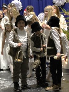 Kilku chłopców w strojach pasterzy stoi na scenie, trzymając latarenkę i rekwizyty. Za nimi znajduje się grupa dzieci w białych strojach aniołów oraz świąteczne dekoracje.