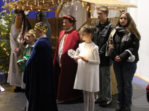 Dzieci w strojach Trzech Króli oraz inni uczestnicy stoją obok szopki bożonarodzeniowej. Obok Maryi i Józefa znajduje się dziewczynka w białej sukience trzymająca świecący rekwizyt.