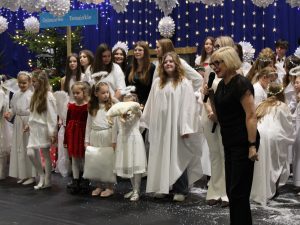 Grupa dzieci w strojach aniołów stoi na scenie, a pani dyrektor z mikrofonem przemawia. Wśród dzieci znajduje się uczestniczka w czerwonej sukience oraz dziecko trzymające pluszową owieczkę