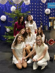 Grupa dziewcząt w białych strojach pozuje na tle świątecznych dekoracji i choinki. Z przodu klęczą trzy dziewczęta, obok nich dorosła opiekunka.