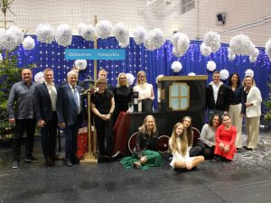 Grupa dorosłych i młodzieży pozuje razem na scenie udekorowanej świątecznie. W tle papierowe ozdoby, niebieska kurtyna z lampkami oraz dekoracja stylizowana na pociąg z tabliczką ‘Ostaszewo – Toruńskie’.