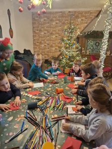 Dzieci siedzą przy stole i rysują kartki świąteczne, używając kredek i flamastrów.