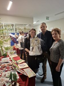 Grupa osób pozuje przy stoisku kiermaszu bożonarodzeniowego, trzymając zapakowaną figurkę anioła. W tle widoczne dekoracje świąteczne.