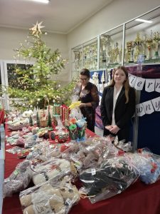 Dwie osoby stoją za stołem kiermaszowym z zapakowanym rękodziełem świątecznym. W tle choinka i baner kiermaszu.