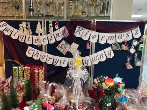 Stoisko kiermaszu bożonarodzeniowego w szkole z rękodziełem i ozdobami świątecznymi. Nad stołem widoczny baner z napisem „Świąteczny Kiermasz Charytatywny”.