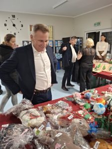 Osoba stojąca przy stole kiermaszowym ogląda zapakowane rękodzieło i ozdoby bożonarodzeniowe wystawione na sprzedaż.