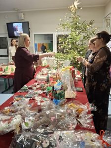 Uczestnicy szkolnego kiermaszu bożonarodzeniowego oglądają i kupują świąteczne dekoracje na stoisku. W tle choinka ozdobiona lampkami.