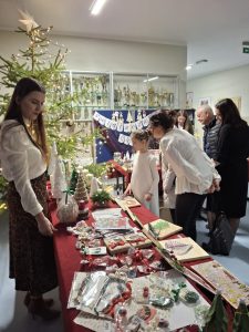 Uczestnicy szkolnego kiermaszu bożonarodzeniowego oglądają rękodzieło świąteczne na stoisku. Obok stoi udekorowana choinka.