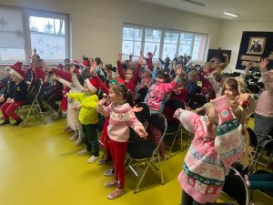 Dzieci z klas 0–3 stoją przy krzesłach w jasnej sali i wykonują ruchy rękami zgodnie z poleceniami. Wiele z nich ma świąteczne stroje i czerwone czapki. Na oknach widać papierowe dekoracje w kształcie śnieżynek.