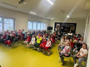 Dzieci siedzą na krzesłach ustawionych w rzędach i oglądają spektakl. Na ścianie w tle znajduje się tablica z napisem informującym o konkursie plastyczno-recytatorskim. Większość dzieci ma świąteczne elementy strojów.