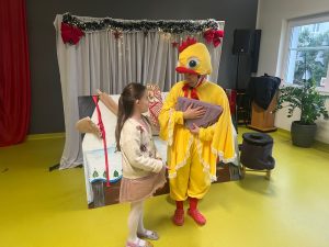 Dziecko rozmawia z aktorem przebranym za żółte pisklę, który trzyma poduszkę będącą elementem sceny. Za nimi znajduje się scenografia kuchni z mąką, rodzynkami i naczyniami.