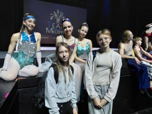 Dwie uczennice klasy siódmej pozuje przed sceną musicalu Kosmos. W tle siedzą trzy tancerki w kolorowych kostiumach, a z prawej strony widoczni są inni artyści odpoczywający po występie.