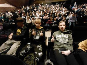 Dwaj uczniowie klasy siódmej siedzą w fotelach widowni CKK Jordanki, pokazując uniesione kciuki. W tle widoczna jest duża publiczność oczekująca na rozpoczęcie musicalu.