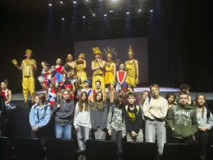 Uczniowie pozują grupowo pod sceną po zakończonym musicalu Kosmos. Na scenie stoją i siedzą wykonawcy w złotych kostiumach oraz w strojach inspirowanych tematyką kosmiczną. Tło stanowi oświetlona scena z logo teatru.