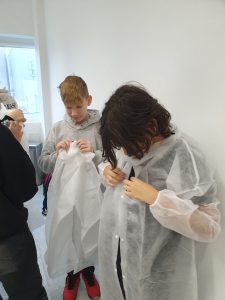 Dwóch uczniów przygotowujących się do zwiedzania zakładu, zakładających jednorazowe fartuchy ochronne w pomieszczeniu.