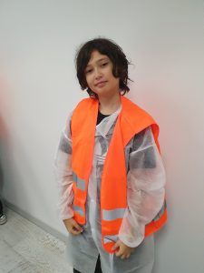 Uczeń ubrany w fartuch ochronny i kamizelkę odblaskową stoi w pomieszczeniu zakładu przed rozpoczęciem zwiedzania.