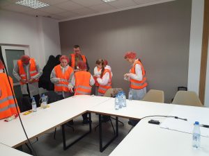 Uczniowie i opiekunowie w kamizelkach odblaskowych i czepkach ochronnych w sali szkoleniowej, przygotowujący się do wejścia na teren produkcji.