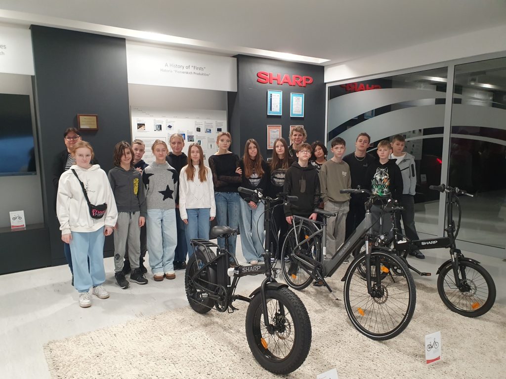 Grupa uczniów klasy VII wraz z opiekunami stoi w holu firmy Sharp. W tle widoczne jest logo firmy oraz plansze informacyjne. Na pierwszym planie znajdują się trzy rowery elektryczne marki Sharp.