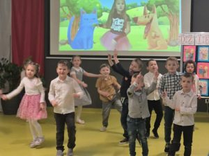 Dzieci tańczą i wykonują ruchy do muzyki w sali przedszkolnej; w tle widoczny ekran z animacją.