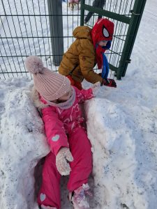 Dzieci bawią się w śnieżnym tunelu; jedno dziecko siedzi w wykopanym przejściu, inne stoi obok i pomaga w zabawie.