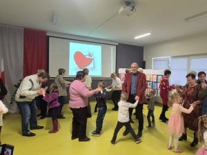 Dzieci i dorośli tańczą parami w sali przedszkolnej; na ekranie w tle wyświetlona jest grafika z sercem.