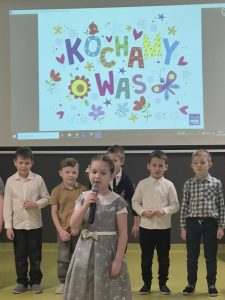 Dziewczynka z mikrofonem stoi przed grupą dzieci, na ekranie za nimi wyświetlony jest napis „Kochamy Was”.