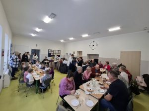 Widok ogólny sali przedszkolnej podczas spotkania z okazji Dnia Babci i Dziadka; dzieci i seniorzy siedzą przy stołach i rozmawiają.