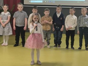 Dziewczynka stoi na środku sali i trzyma mikrofon, za nią ustawiona jest grupa dzieci przygotowujących się do występu.