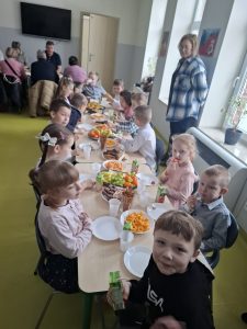 Dzieci siedzą przy długim stole i spożywają poczęstunek w sali przedszkolnej; w tle widoczna nauczycielka oraz opiekunowie.