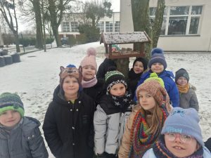 Dzieci z oddziału przedszkolnego stoją na zewnątrz przy karmniku dla ptaków zawieszonym na drzewie.