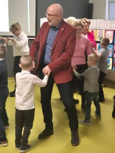 Dorosły mężczyzna trzyma za ręce dziecko i tańczy z nim w sali przedszkolnej; w tle widoczni inni dorośli i dzieci.