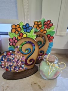 Prace plastyczne wykonane przez dzieci – dekoracyjne serca z elementami kwiatowymi i koralikami oraz mały słoiczek z kolorową solą, ustawione na parapecie.