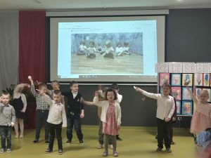 Grupa dzieci stoi na środku sali z uniesionymi rękami podczas występu; w tle widoczny ekran i tablica z pracami plastycznymi.