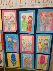 Tablica z pracami plastycznymi dzieci przedstawiającymi babcie i dziadków, narysowanymi kredkami i umieszczonymi na kolorowych kartkach.