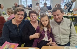 Dziewczynka siedzi przy stole pomiędzy swoimi dwiema babciami i dziadkiem; wszyscy pozują do zdjęcia podczas przedszkolnego spotkania.