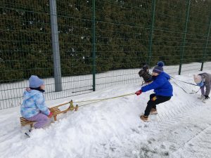 Dzieci z oddziału przedszkolnego bawią się na śniegu, ciągnąc sanki po zaśnieżonej nawierzchni w pobliżu ogrodzenia.