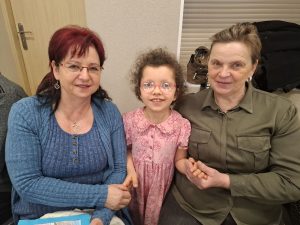 Dziewczynka stoi pomiędzy dwiema kobietami, trzymając je za ręce; pozują do zdjęcia w sali przedszkolnej.