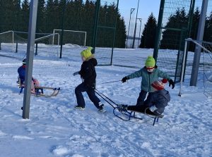 Kilka dzieci bawi się na śniegu, ciągnąc sanki w parach na ogrodzonym terenie rekreacyjnym.