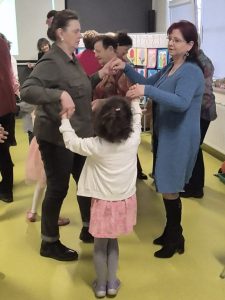 Dorosłe kobiety i dziecko trzymają się za ręce i tańczą w sali przedszkolnej podczas wspólnej zabawy.