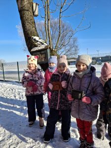 Grupa dzieci w zimowych ubraniach stoi na śniegu przy drzewie z karmnikiem; część dzieci trzyma małe aparaty fotograficzne.