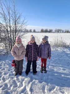 Trójka dzieci ubranych w zimowe kurtki i czapki stoi na zaśnieżonym polu; w tle widoczne drzewa i jasne, bezchmurne niebo.